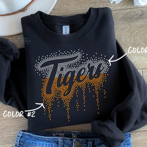 May include: Black sweatshirt with the word "Tiger" in white and orange rhinestones. The word "Tiger" is in a script font with a dripping effect. The sweatshirt is folded on top of blue jeans.