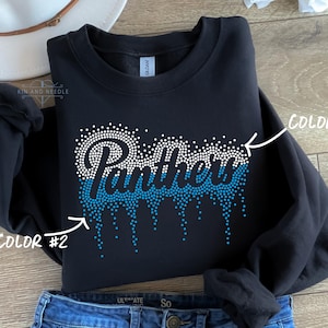 May include: Black sweatshirt with the word "Panthers" in white and blue rhinestone design. The word is stylized with a dripping effect. The sweatshirt is folded on top of blue jeans.