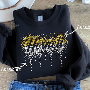 May include: Black crewneck sweatshirt with the word "Hornets" in yellow and white rhinestone design. The word is stylized with a dripping effect. The sweatshirt is folded on top of blue jeans.
