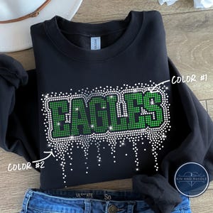 May include: Black crewneck sweatshirt with the word "EAGLES" in green and white rhinestones. The design features a dripping rhinestone effect. The sweatshirt is folded on top of blue denim jeans.