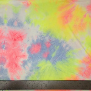 May include: A white fabric with a tie-dye pattern in shades of pink, blue, yellow, and green. The fabric is shown with a ruler for scale.