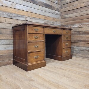 May include: An antique wooden desk with multiple drawers and brass handles. The desk has a warm, brown finish and is set against a backdrop of wooden paneling. The desk has a classic design, with a rectangular shape and a spacious work surface.