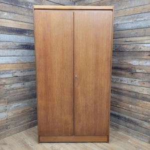 May include: A tall, rectangular wooden wardrobe featuring two full-length doors with a smooth, light brown wood grain finish. The cabinet has a simple, clean-lined design with a single small metal keyhole centred between the two doors.
