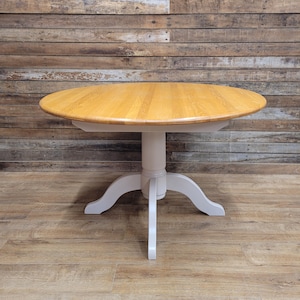 May include: A round dining table featuring a natural light brown wood grain top and a solid light grey pedestal base with four curved legs extending outward to provide support for the circular tabletop surface.