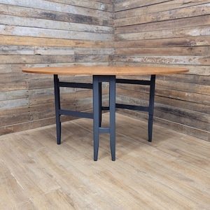 May include: A round wooden table with a light brown tabletop and dark blue legs. The table is approximately 122 cm in diameter when fully extended. The table is set against a wooden plank wall.