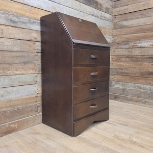 May include: A dark brown wooden desk with four drawers and a slanted top. The desk has a small handle on the top and a decorative trim above the drawers. The desk is made of wood and has a vintage appearance.