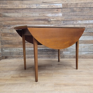May include: A mid-century modern, wooden drop-leaf table with a warm brown finish. The table has a semi-circular shape when the leaf is down and stands on four slender, tapered legs. The table's dimensions are approximately 76 cm long, 38 cm wide, and 71 cm high.