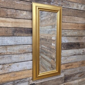 May include: A long, rectangular mirror with a gold frame. The frame has a detailed design. The mirror reflects the wooden wall behind it, made of horizontal planks in shades of brown and grey. The mirror is a decorative item.