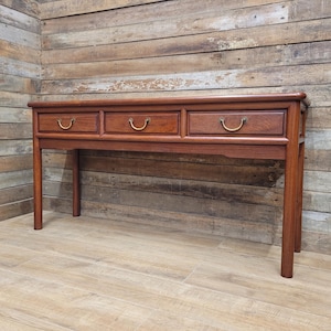 May include: A dark brown wooden console table with three drawers and brass-coloured handles. The table has a rectangular top and four slender legs. The background is a wooden wall and floor.
