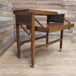 May include: A vintage wooden side table with a fold-out top and caster wheels. The table is made from dark wood and has a rectangular shape with a pull-out drawer. The table's design is simple and functional, suitable for various uses.