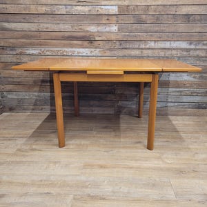 May include: A rectangular light brown wooden dining table with four straight legs and two extendable side leaves. The table features a smooth, flat surface with a visible wood grain pattern and a horizontal support frame connecting the four legs.