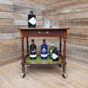 May include: A dark brown wooden drinks trolley with a green lower shelf and four bottles of spirits. The trolley has a drawer and is on wheels. Two clear glasses and a bottle of gin are on the top shelf.