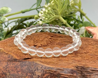 Peut inclure: Un bracelet en perles de cristal de roche clair sur une surface en bois avec de l'herbe verte au premier plan. Le bracelet est composé de petites perles rondes.