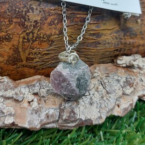 May include: A silver chain necklace with a pink and grey ruby pendant. The pendant is a raw, uncut stone. The necklace is on a wooden surface with green grass in the background. The card attached to the necklace reads "Energy Crystals Handmade Jewellery Ruby Attributes: Energy, Leading Abilities Sexual Activity, Concentration Sociability, Passion Therapy: Detox Body, Blood Lympho, Lungs Reproductive Organs Blood's Flow jpmineralstone.com"