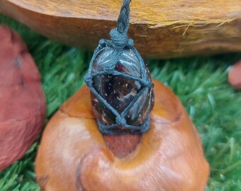 May include: A dark red gemstone wrapped in black cord, hanging from a black cord necklace. The gemstone is tied with a knot at the top and bottom.