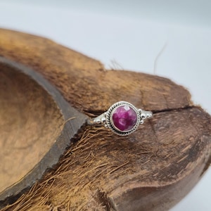 Peut inclure: Une bague en argent ornée d'une pierre précieuse ronde rose foncé. La bague a un anneau détaillé et texturé et un sertissage décoratif autour de la pierre. La bague est présentée sur une surface brune et texturée.