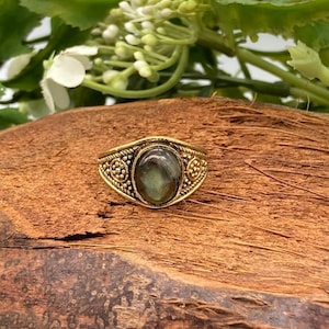 Pode incluir: Um anel de ouro com uma gema verde escura engastada em uma banda decorativa. O anel está sobre um pedaço de madeira.