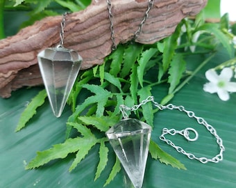 May include: Two clear quartz crystal pendulums with silver chains hanging from a piece of driftwood on a green leaf background.