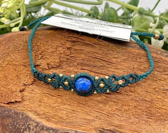 Puede incluir: Una pulsera de macramé hecha a mano con una vibrante piedra preciosa azul en el centro. El cordón verde oscuro está acentuado con pequeñas cuentas doradas. La pulsera se exhibe sobre una superficie de madera con una etiqueta adjunta.
