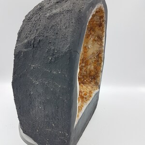 May include: A large, dark gray geode with a natural opening revealing a cluster of small, light brown crystals. The geode is sitting on a clear, round base.