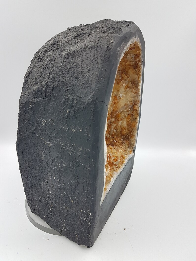 May include: A large, dark gray geode with a natural opening revealing a cluster of small, light brown crystals. The geode is sitting on a clear, round base.