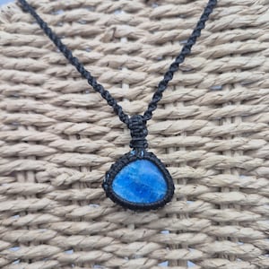 May include: A handmade necklace featuring a vibrant blue gemstone pendant. The pendant is encased in a black macrame setting and suspended from a matching black cord. The necklace is displayed against a textured, woven background.