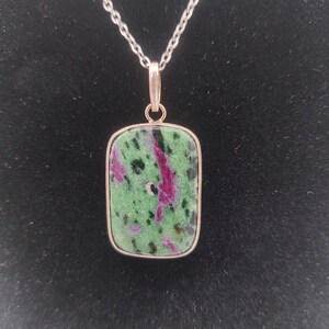 Op de afbeelding: Een zilveren ketting met een rechthoekige hanger met een groene en roze edelsteen. De edelsteen heeft een gevlekt patroon in tinten groen, roze en zwart.