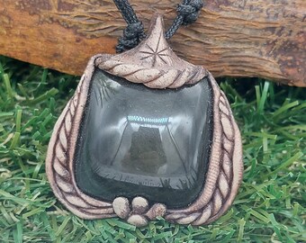 May include: A handcrafted pendant necklace featuring a dark, square stone set within a decorative, light brown frame. The frame has a leaf-like design and is suspended from a black cord. The necklace rests on a bed of green grass.