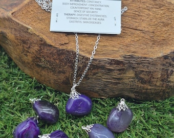 Puede incluir: Un collar de cadena plateada con un colgante grande de ágata púrpura pulida. Varias piedras de ágata sueltas en diferentes tonos de púrpura están dispersas alrededor del collar. Una etiqueta con el texto "ENERGY CRYSTALS handmade" y atributos de ágata es visible.