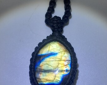May include: A black cord necklace with a large oval-shaped labradorite gemstone pendant. The gemstone has a blue, yellow, and black iridescent sheen.
