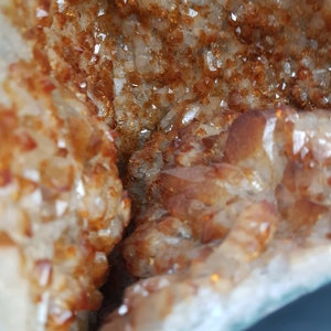 May include: Close-up of a natural crystal geode with a rough, textured exterior and a sparkling, translucent interior. The geode is filled with small, orange-brown crystals.