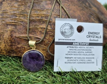 May include: A gold chain necklace with a round amethyst pendant. The pendant is set in a gold bezel and has a purple hue. The necklace is on a green grassy surface with a white card that reads "ENERGY CRYSTALS handmade AMETHYST ATTRIBUTES: MENTAL SECURITY. MIND RELAXATION MAKING DECISIONS, MENTAL SUPPORT, MEMORY BOOSTER, VISUALIZATION THERAPY: METABOLISM, HEADACHE GEOPATHETIC STRESS, LUNGS, RESPIRATORY, HORMONES BOOSTER"