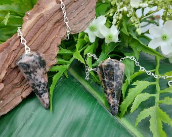 May include: Two pink and black crystal pendulums with silver chains hanging on a green leaf and brown wood background.