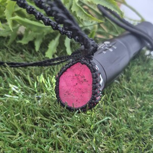 Puede incluir: Una linterna negra con un colgante de piedra rosa, suspendido por un cordón de macramé negro. El colgante es de un color rosa vibrante y está encerrado en un diseño tejido negro. La linterna está sobre un lecho de césped artificial verde.