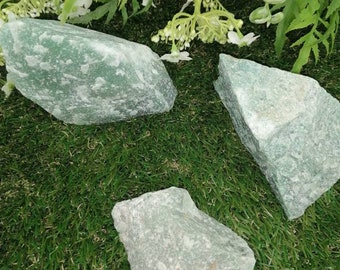 May include: Three green aventurine crystal stones on a green artificial grass background. The stones are rough and have a natural, unpolished look.