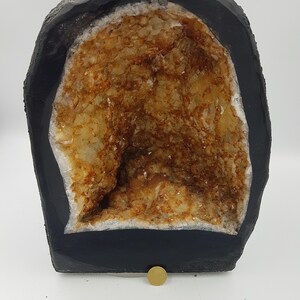May include: A large geode with a black exterior and a hollow interior filled with clear quartz crystals with a brown tint. The geode is sitting on a white surface with a ruler for scale.