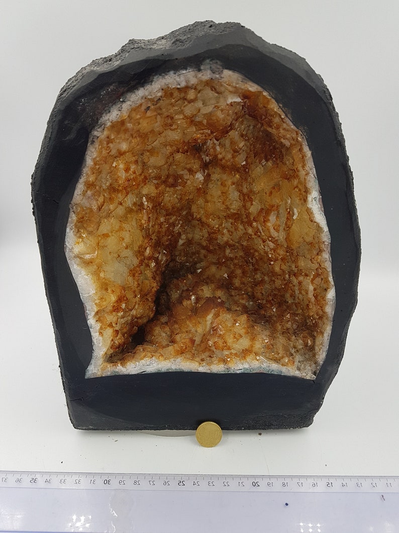 May include: A large geode with a black exterior and a hollow interior filled with clear quartz crystals with a brown tint. The geode is sitting on a white surface with a ruler for scale.
