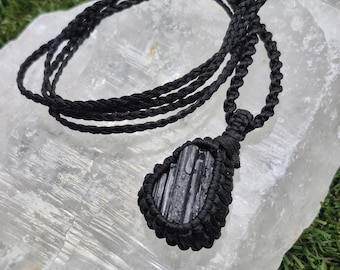 May include: A black macrame necklace featuring a dark, rectangular crystal pendant. The pendant is encased in a woven black cord, and the necklace has a long, braided cord. The necklace is displayed on a white, textured surface.