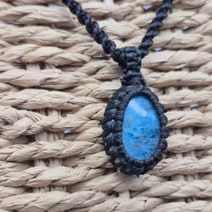 May include: A handmade macrame necklace featuring a blue gemstone pendant. The pendant is oval-shaped and encased in black macrame. The necklace cord is also black, with a woven texture. The background is a woven, light brown material.