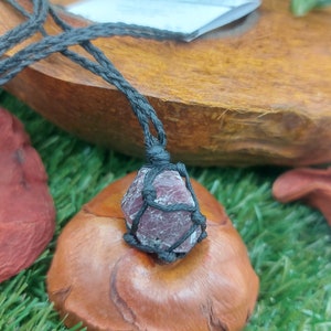 May include: A dark red gemstone wrapped in black cord, hanging from a black cord necklace. The gemstone is on a wooden surface.