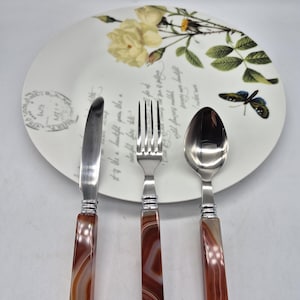 Puede incluir: Un juego de tres cubiertos de plata con mangos de ágata. El cuchillo, el tenedor y la cuchara tienen mangos de ágata marrón y blanco.