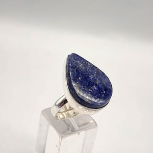 May include: A silver ring featuring a large, teardrop-shaped lapis lazuli stone. The deep blue stone has flecks of gold and is set in a simple, polished silver band. The ring is displayed on a clear, rectangular stand.