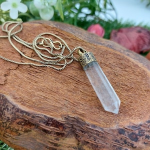 Peut inclure: Un pendentif en cristal de quartz transparent avec un sertissage en métal doré orné est suspendu à une chaîne dorée. Le pendentif repose sur un morceau de bois.