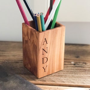 May include: A wooden pencil holder with the name "ANDY" engraved on the front. The holder is filled with colored pencils, including red, green, blue, and yellow.