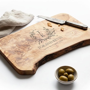 May include: A wooden serving board with a natural edge, engraved with floral details and the text "TOR AND KEVIN 16TH OCTOBER 2023." A cheese knife and a small bowl of olives are also present. A linen napkin is in the background.