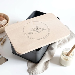 May include: A wooden bread bin with a lid engraved with "Grandma's Homemade Bread" and a graphic. A loaf of bread is in a woven basket. A metal scraper and a jar with a wooden spoon are also visible. The bread bin is grey.