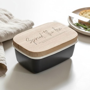 May include: A rectangular butter dish with a wooden lid. The lid is engraved with the words "Spread the love" in a cursive font. The dish is black with a white rim. A piece of bread and a knife are in the background.