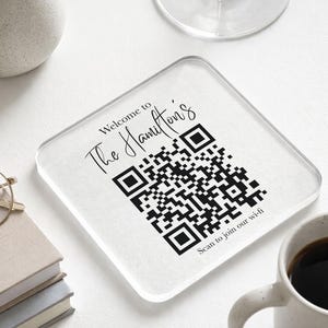 May include: A clear, square coaster featuring the text "Welcome to The Hamilton's" and a QR code. The words "Scan to join our wi-fi" are below the QR code. A coffee mug and other items are in the background.