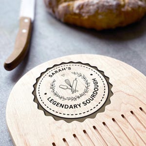 Pu&ograve; includere: Un tagliere di legno chiaro con un emblema decorativo rotondo che recita "Sarah's Legendary Sourdough". L'emblema presenta una frusta, un mattarello e un cucchiaio all'interno di una corona d'alloro. Un coltello da pane &egrave; appoggiato sul tagliere.