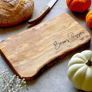 May include: A wooden cutting board with a natural edge, engraved with "Brian's Pizzeria HAPPY 50TH". A bread knife and pumpkins are nearby. The board is made of light brown wood.